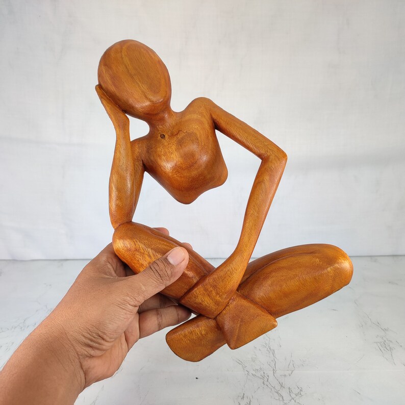 May include: A wooden sculpture of a person sitting with their head resting on their hand. The sculpture is made of light brown wood and has a smooth finish.