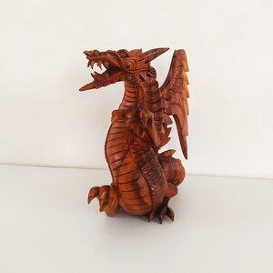 May include: A wooden dragon sculpture with intricate details, carved in a reddish-brown colour. The dragon is depicted with wings spread, a fierce expression, and a curled tail.