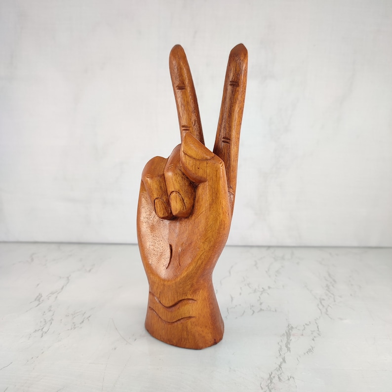 May include: A wooden hand sculpture in a peace sign gesture. The hand is carved from light brown wood and has a detailed finish.