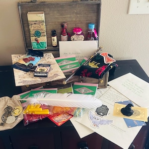 May include: A wooden trunk filled with fantasy-themed items including a white feather quill, small glass bottles, a black wand, a burlap pouch with metal coins, envelopes addressed to Mr. H. Potter, a chocolate bar, and various stickers featuring an owl and a hat.