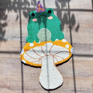 May include: A whimsical, handmade rug in the shape of a frog sitting on a mushroom. The frog is green with pink cheeks and a purple party hat. The mushroom has a white stem and a yellow and white cap. The rug is placed on a wooden floor.