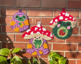 May include: Three handmade mushroom-shaped wall hangings with embroidered frog faces. The mushrooms are purple, green, and beige with white polka dots. The mushrooms are hanging on a brick wall.