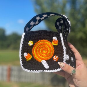 May include: A black crocheted bag with a white and black patterned strap. The bag features a colorful candy lollipop and candy corn designs.