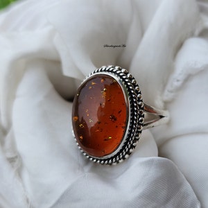 May include: A silver ring featuring a large oval amber stone set in a beaded bezel. The stone has a deep orange colour with flecks of gold.
