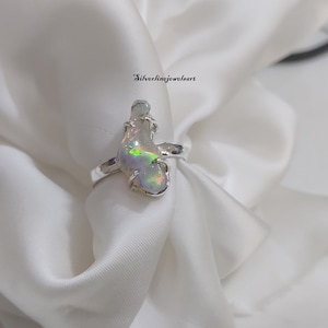 May include: A silver ring with a large, iridescent opal stone set in a wire-wrapped design. The ring is displayed on a white fabric background.