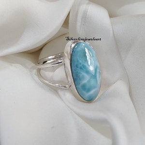 May include: A silver ring featuring a light blue oval-shaped gemstone. The gemstone has a swirling pattern and a smooth, polished finish. The ring is displayed on a white fabric background.