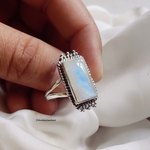 May include: A silver ring with a rectangular moonstone set in a detailed silver bezel. The ring is on a finger.