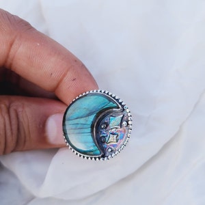 Könnte beinhalten: Ein runder Silberring mit einem großen, irisierenden blau-grünen Stein. Der Stein hat eine Halbmondform mit einem dekorativen Design an der Seite. Der Ring ist auf weißem Hintergrund platziert.