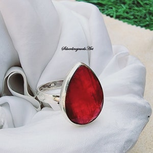 May include: A silver ring with a large, teardrop-shaped, red gemstone. The ring is on a white fabric background.