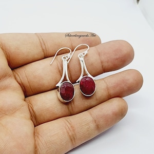 Könnte beinhalten: Ein Paar silberne Ohrhänger mit roten Rubin-Edelsteinen. Die Ohrringe haben ein schlichtes, elegantes Design mit einem geschwungenen, durchbrochenen Silberteil oben und einem runden Rubin-Edelstein unten.