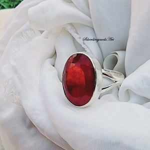 May include: A silver ring with a large, oval-cut, deep red gemstone. The gemstone is faceted, catching the light and displaying a brilliant shine. The ring band splits into two strands on either side of the stone. The ring is set against a white fabric.