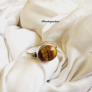 May include: A silver ring with a round, brown and gold tiger's eye gemstone. The ring has a textured band and three small silver balls on one side.