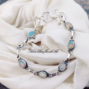 Puede incluir: Un brazalete de plata con cierre de palanca y cinco piedras de ópalo de forma ovalada. El brazalete está sobre un fondo de tela blanca.