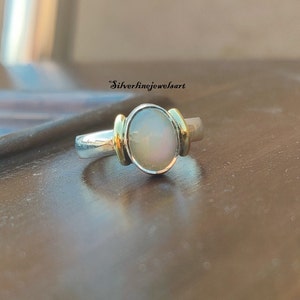 May include: A silver ring with a white oval gemstone set in a gold bezel. The ring has a simple band.