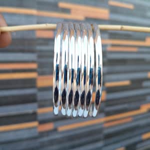 Könnte beinhalten: Eine Sammlung glänzender silberner Armreifen, die das Licht reflektieren, wird auf einem dünnen Holzstab präsentiert. Die Armreifen sind gestapelt und zeigen ihre glatte, polierte Oberfläche. Der Hintergrund zeigt eine strukturierte Wand mit grauen und orangefarbenen rechteckigen Mustern.