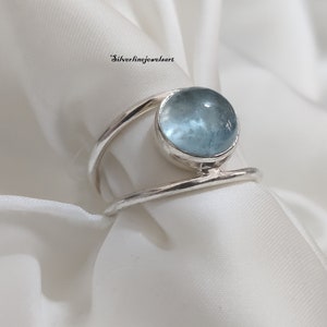 Puede incluir: Un anillo de plata con una piedra preciosa de aguamarina azul claro. El anillo tiene un diseño simple y moderno con una banda delgada que envuelve la piedra.