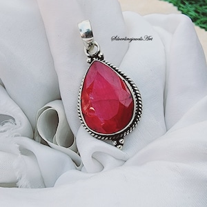 Könnte beinhalten: Ein tropfenförmiger Anhänger mit einem roten Edelstein, der in einen silbernen Rahmen gefasst ist. Der Anhänger liegt auf einem weißen Stoffhintergrund. Der Text "Silverlingjewels.Art" ist in der oberen rechten Ecke des Bildes sichtbar.