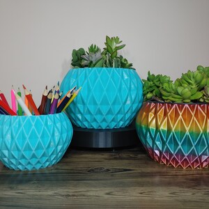 May include: Three decorative, geometric-patterned planters. One is filled with coloured pencils, another with succulents, and the third with succulents. The planters are turquoise and rainbow-coloured, sitting on a wooden surface.