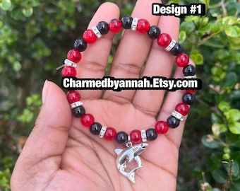 May include: Red and black beaded bracelet with a silver shark charm. The bracelet features red and black beads, interspersed with silver spacers. The charm is a detailed shark pendant. The text "Design #1" and "Charmedbyannah.Etsy.com" are visible.