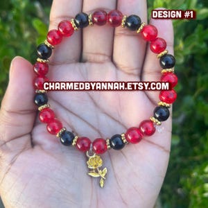 May include: A beaded bracelet featuring red and black beads with gold accents. The bracelet has a gold rose charm and the text "DESIGN #1" and "CHARMEDBYANNAH.ETSY.COM". The bracelet is likely handmade.