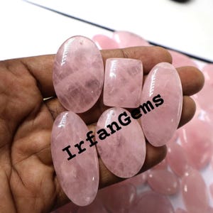 May include: A collection of polished rose quartz gemstones in various shapes, including oval and rectangular. The stones are a soft pink color and have a translucent quality. The text "IrfanGems" is printed on the stones.