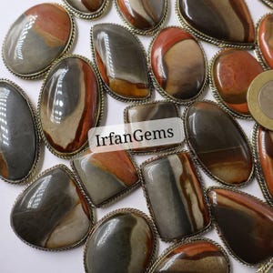 Può includere: Una collezione di cabochon di pietre preziose lucide in varie forme, tra cui ovali, a goccia e rettangolari. Le pietre presentano una miscela di tonalità terrose come marrone, grigio e arancione, incastonate in castoni di metallo color argento. Il testo "IrfanGems" è visibile.