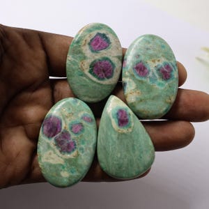 May include: Four polished gemstones with a green matrix and vibrant red inclusions. The stones vary in shape, including oval and teardrop, and are held in a hand against a white background. The gemstones have a natural, organic appearance.