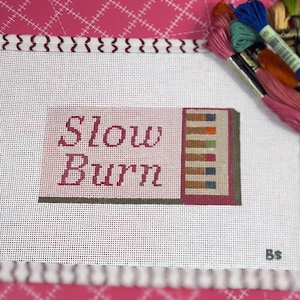 May include: A needlepoint canvas with the words "Slow Burn" in burgundy script. The canvas features a small, colorful geometric design and is surrounded by spools of embroidery floss in various colors, including pink, blue, and orange.