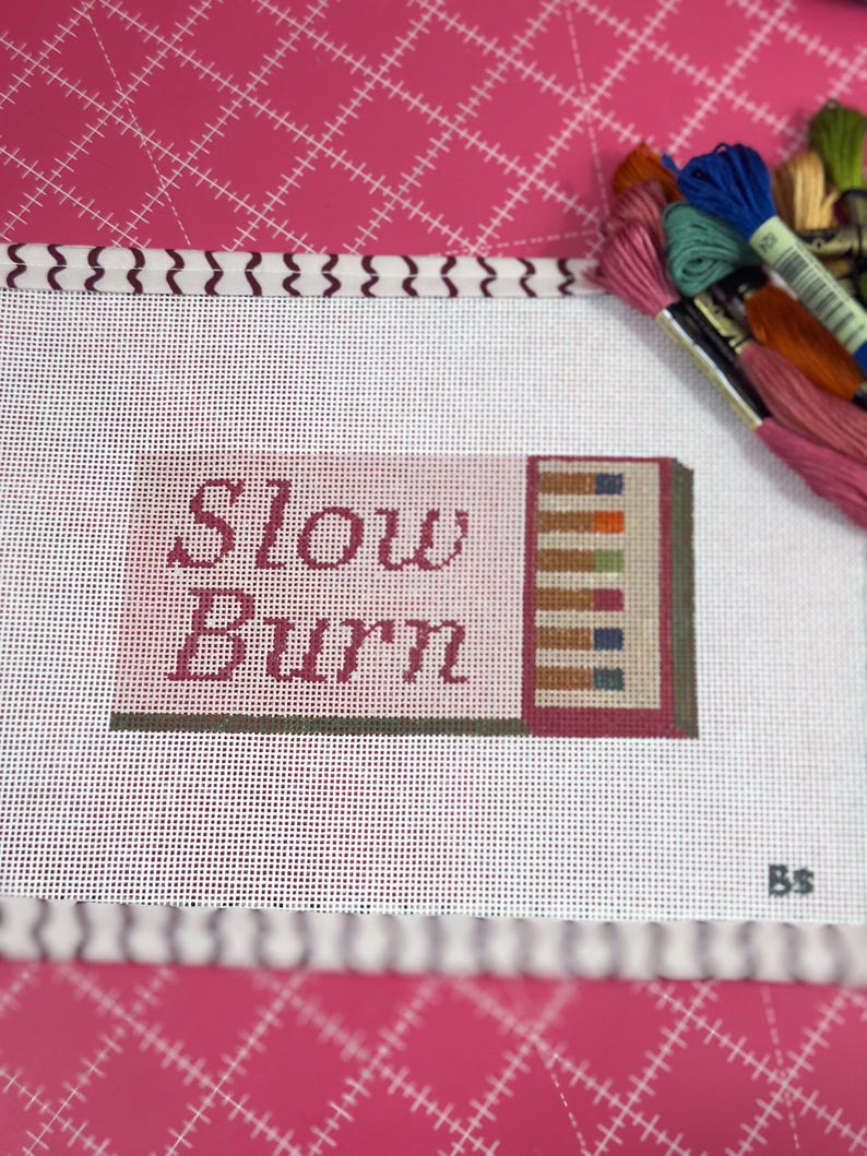 May include: A needlepoint canvas with the words "Slow Burn" in burgundy script. The canvas features a small, colorful geometric design and is surrounded by spools of embroidery floss in various colors, including pink, blue, and orange.