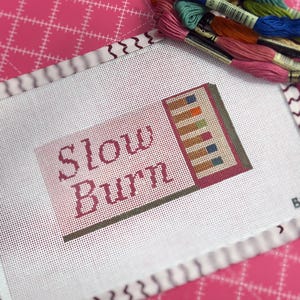 May include: A needlepoint canvas with the words "Slow Burn" in burgundy thread. The canvas features a stitched design of colorful rectangles and is surrounded by a white border. A collection of embroidery floss in various colors rests on a pink cutting mat.