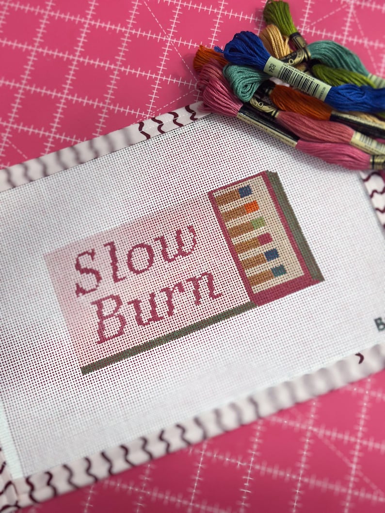 May include: A needlepoint canvas with the words "Slow Burn" in burgundy thread. The canvas features a stitched design of colorful rectangles and is surrounded by a white border. A collection of embroidery floss in various colors rests on a pink cutting mat.