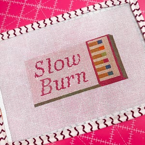 May include: Needlepoint canvas with the words "Slow Burn" in pink script. The canvas features a colorful design of stacked books and a patterned border. The background is a bright pink cutting mat.