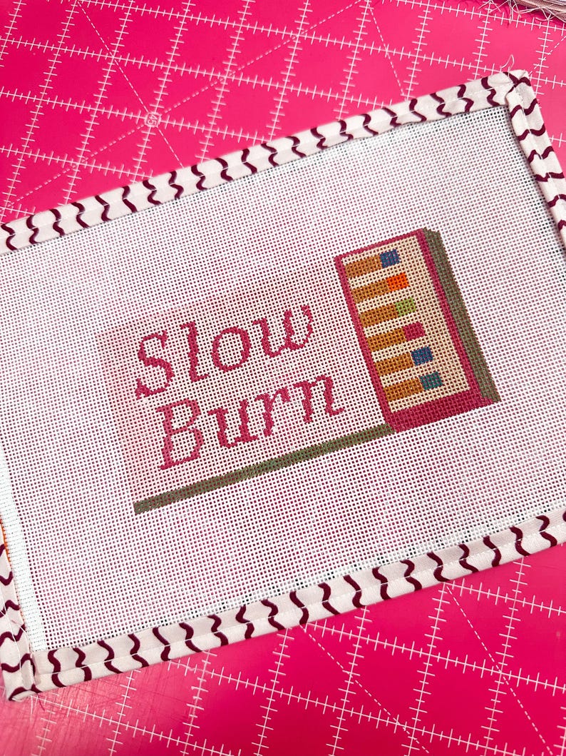 May include: Needlepoint canvas with the words "Slow Burn" in pink script. The canvas features a colorful design of stacked books and a patterned border. The background is a bright pink cutting mat.