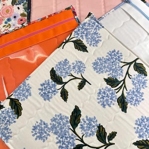 May include: Close-up of several fabric pouches with zippers. One pouch features a white background with blue hydrangea floral print. Other pouches are solid colors: pink, orange, and floral patterns. The pouches are quilted.