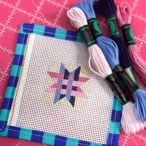 May include: Needlepoint canvas with a geometric star design in pink, blue, and purple. The canvas is framed with a blue and turquoise plaid fabric. Several skeins of embroidery floss in coordinating colors are arranged nearby.