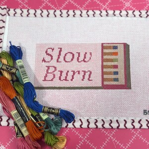 May include: A needlepoint canvas with the words "Slow Burn" in pink lettering. The design includes a stack of books with colorful spines. Several skeins of embroidery floss in various colors are arranged around the canvas, ready for stitching.