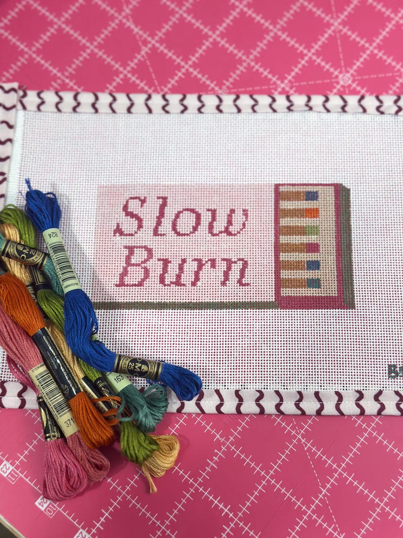 May include: A needlepoint canvas with the words "Slow Burn" in pink lettering. The design includes a stack of books with colorful spines. Several skeins of embroidery floss in various colors are arranged around the canvas, ready for stitching.
