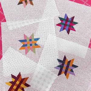May include: Five white needlepoint canvases with colorful star designs. Each star is a different color combination, including brown, pink, blue, orange, green, and purple. The canvases are arranged on a pink background.