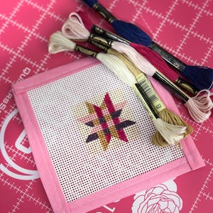 May include: A pink needlepoint project with a star design in shades of pink, purple, and gold. Several skeins of embroidery floss in various colors, including blue, pink, and cream, are arranged around the project. The project is on a pink cutting mat.