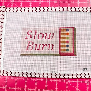 May include: Needlepoint canvas with the words "Slow Burn" in pink script. A small, colorful stack of books is to the right. The canvas is white with a patterned border and is on a pink cutting mat.