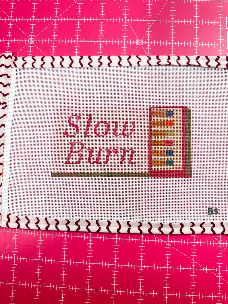May include: Needlepoint canvas with the words "Slow Burn" in pink script. A small, colorful stack of books is to the right. The canvas is white with a patterned border and is on a pink cutting mat.