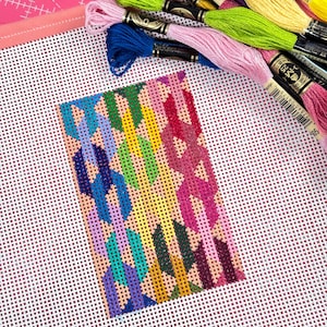 May include: A needlepoint project in progress, featuring a geometric design in various colors including blue, green, pink, and yellow. The canvas is surrounded by spools of colorful embroidery floss in shades of pink, yellow, and green.