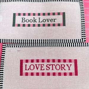 May include: Two needlepoint canvases with black and white striped borders. One canvas reads "Book Lover" in a pink and green rectangular design. The other canvas reads "Love Story" in a pink rectangular design.