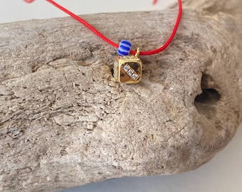 May include: A gold-colored cube pendant necklace with clear rhinestones and a red cord. The necklace features a small blue and white bead. The pendant is resting on a piece of driftwood.
