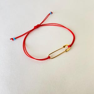 May include: A red string bracelet with a gold safety pin charm. The bracelet is tied with a knot and has two blue beads on the ends.