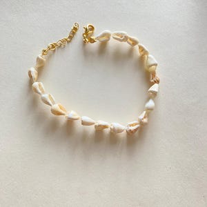 May include: A delicate bracelet featuring small, natural seashells strung together. The shells are primarily off-white with subtle tan markings. The bracelet is secured with a gold-colored clasp and chain extender, adding a touch of elegance to the beach-inspired accessory.