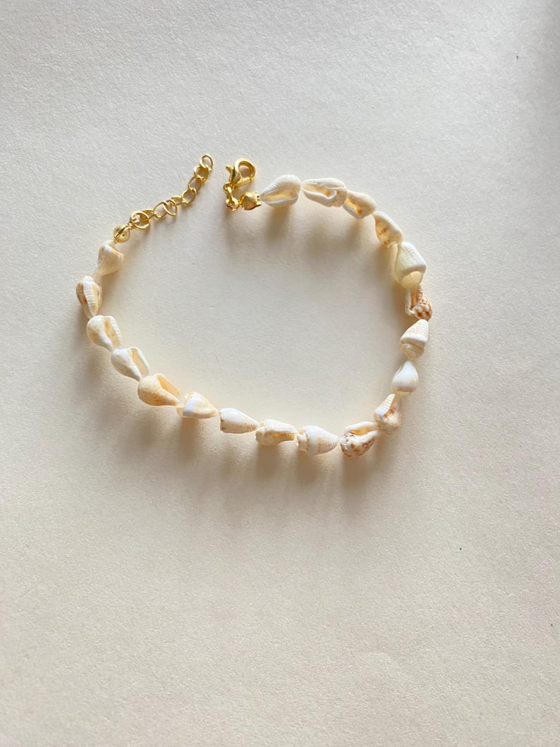 May include: A delicate bracelet featuring small, natural seashells strung together. The shells are primarily off-white with subtle tan markings. The bracelet is secured with a gold-colored clasp and chain extender, adding a touch of elegance to the beach-inspired accessory.