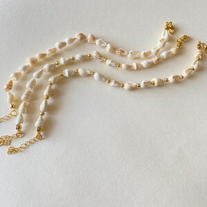 May include: Three seashell necklaces with gold-colored chain and small gold beads. The shells are white and beige, with a natural, organic shape. The necklaces are arranged on a light beige surface.
