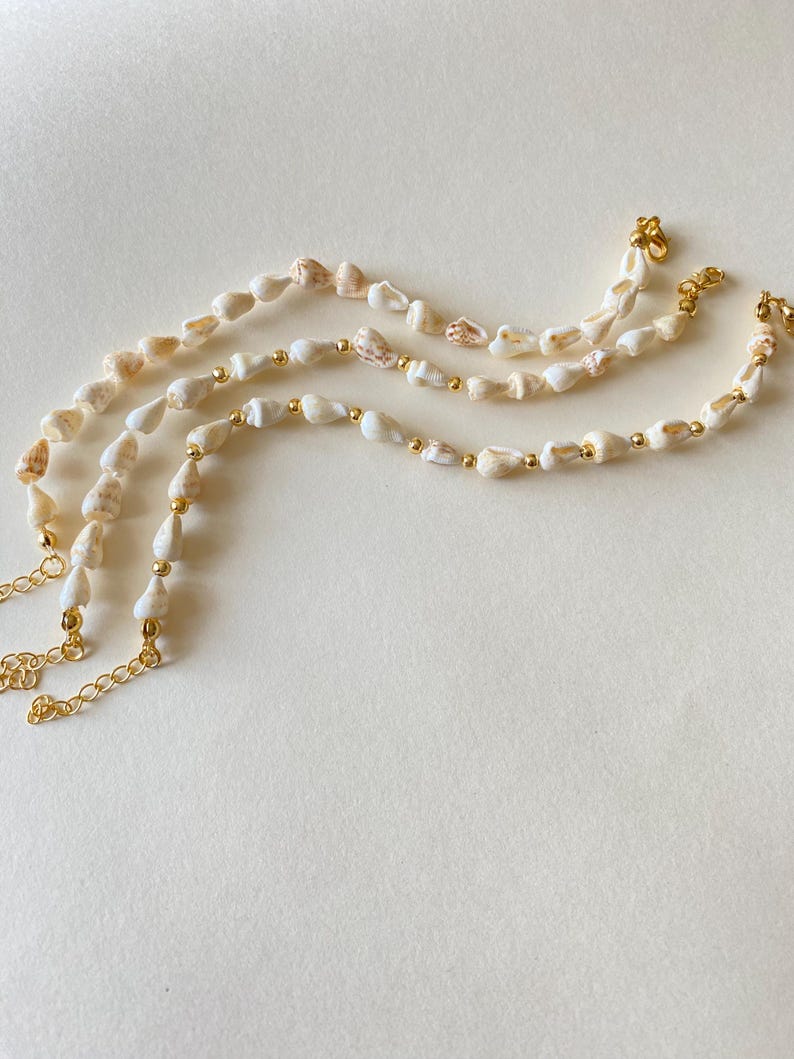 May include: Three seashell necklaces with gold-colored chain and small gold beads. The shells are white and beige, with a natural, organic shape. The necklaces are arranged on a light beige surface.