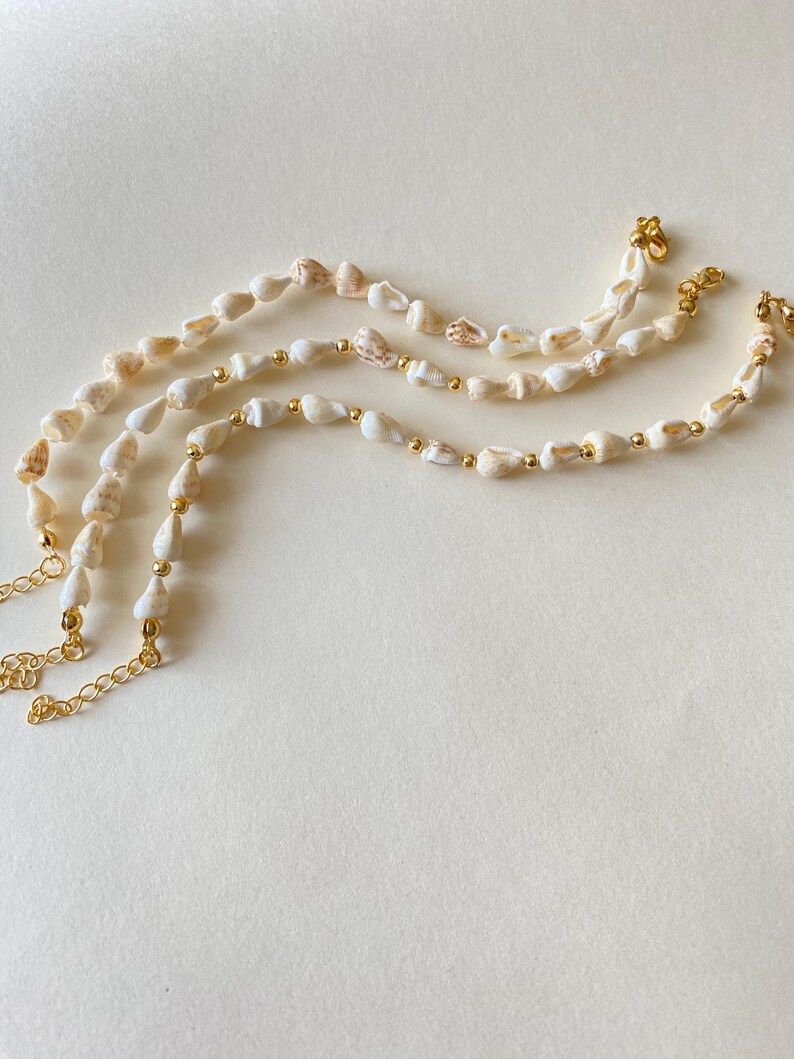 May include: Three delicate necklaces featuring small, natural seashells strung together with gold-colored beads and chains. The seashells are primarily white with subtle tan and brown markings. The necklaces are displayed on a neutral background.